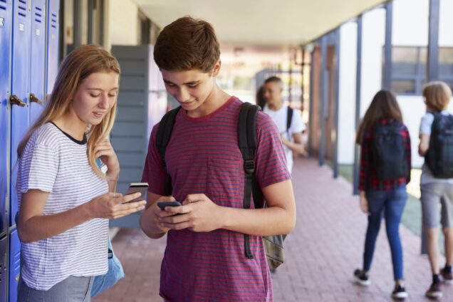 Teenage boy and teenage girl stand by their lockers at school talking and looking at their phones together
