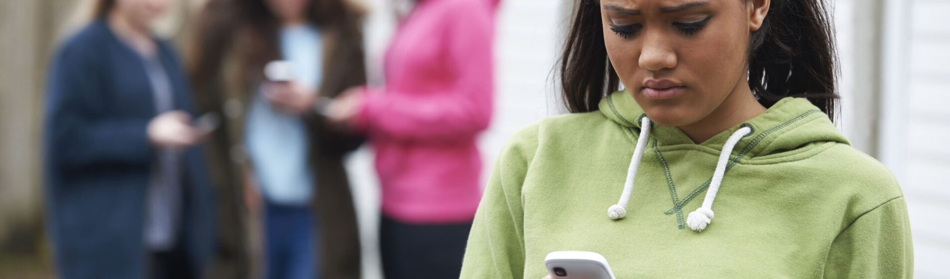 Teenage girl looking worriedly at her phone as three out of focus figures stand behind her all looking at their phones