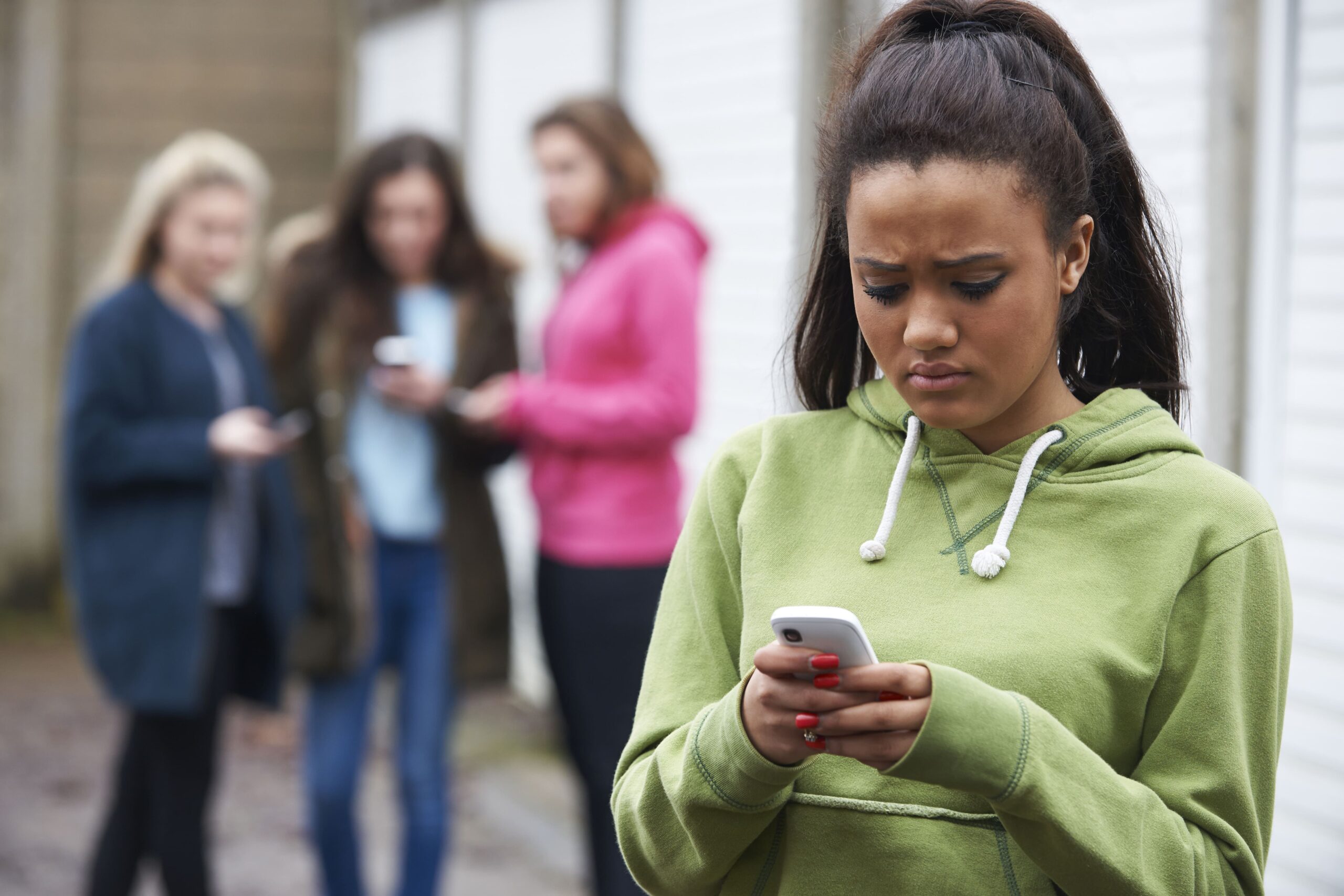 Teenage girl looking worriedly at her phone as three out of focus figures stand behind her all looking at their phones