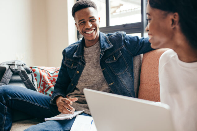 A man and a woman talking together on a sofa, the woman holds a laptop while the man has a notebook and pen
